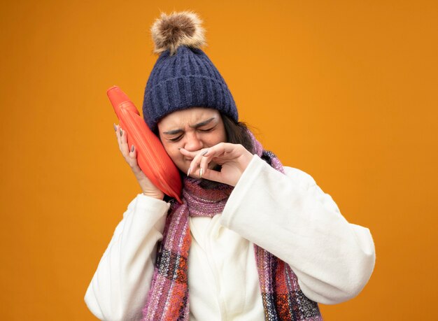 Stirnrunzelnde junge kranke Frau, die Robe Wintermütze und Schal berührt Kopf mit heißem Wasserbeutel abwischend Nase mit geschlossenen Augen lokalisiert auf orange Wand