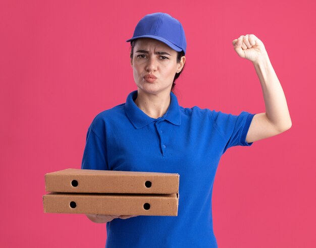 Stirnrunzelnde junge Lieferfrau in Uniform und Mütze, die Pizzapakete hält und nach vorne schaut, die Klopfgeste mit geschürzten Lippen einzeln auf rosa Wand macht?
