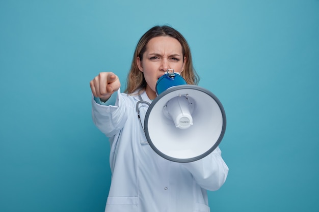 Stirnrunzelnde junge Ärztin, die medizinische Robe und Stethoskop um den Hals trägt, die durch Sprecher sprechen und auf Kamera zeigen