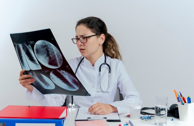 Stirnrunzelnde junge Ärztin, die medizinische Robe und Stethoskop und Brille trägt, sitzt am Schreibtisch mit medizinischen Werkzeugen, die Röntgenaufnahme lokalisieren und betrachten