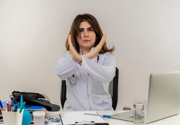 Stirnrunzelnde Ärztin mittleren Alters, die medizinische Robe und Stethoskop trägt, sitzt am Schreibtisch mit Zwischenablage des medizinischen Werkzeugs und Laptop, die keine Geste isoliert tun