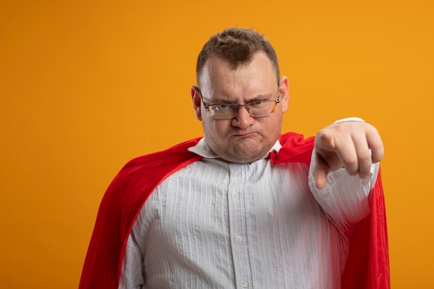 Kostenloses Foto stirnrunzelnder erwachsener slawischer superheldenmann im roten umhang, der die brille trägt, die lokal auf orange wand schaut und zeigt