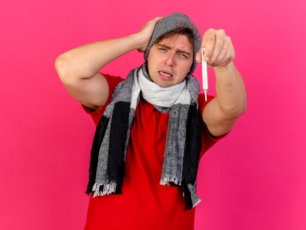 Stirnrunzelnder junger hübscher blonder kranker Mann, der Wintermütze und Schal trägt, die Thermometer nach vorne ausdehnen, Hand auf Kopf halten, die vorne auf rosa Wand lokalisiert betrachtet