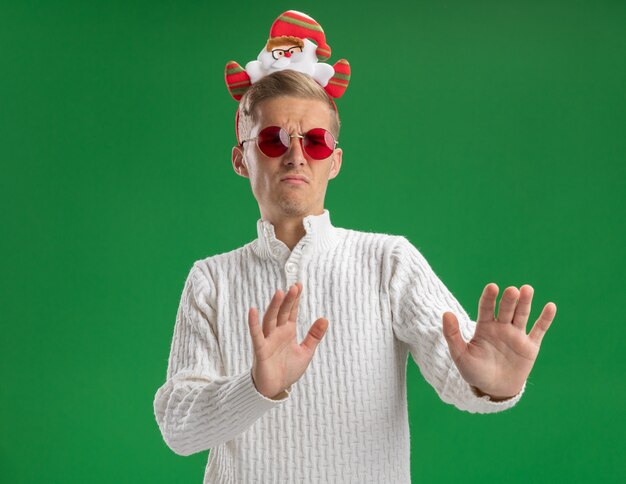 Stirnrunzelnder junger hübscher Kerl, der Weihnachtsmann-Stirnband mit Brille trägt, die Seite betrachtet, die Ablehnungsgeste lokalisiert auf grüner Wand mit Kopienraum tut