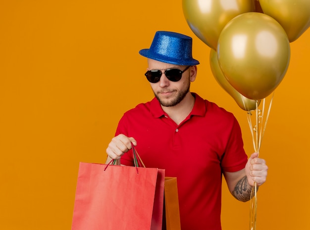 Stirnrunzelnder junger hübscher Party-Typ, der Sonnenbrille und Partyhut hält, die Luftballons und Papiertüten lokalisiert auf orange Wand hält