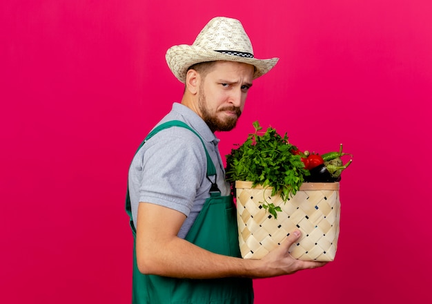 Stirnrunzelnder junger hübscher slawischer Gärtner in Uniform und Hut, die in der Profilansicht stehen und Korb des Gemüses halten