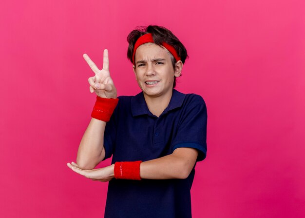 Stirnrunzelnder junger hübscher sportlicher Junge, der Stirnband und Armbänder mit Zahnspangen trägt, die Hand unter Ellbogen setzen, der nach vorne Friedenszeichen lokalisiert auf purpurroter Wand tut