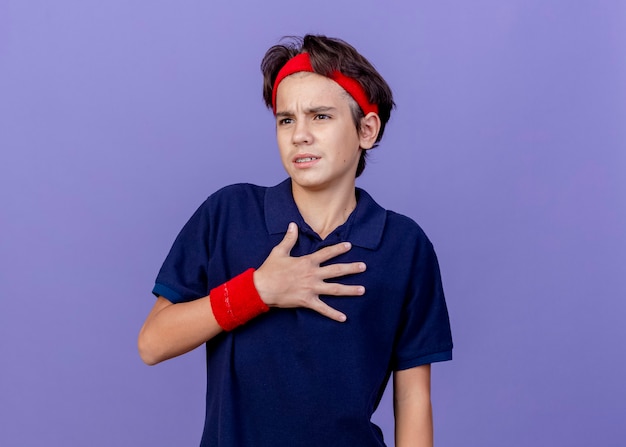 Stirnrunzelnder junger hübscher sportlicher Junge, der Stirnband und Armbänder mit Zahnspangen trägt, die Seite betrachten, die Hand auf Brust lokalisiert auf lila Wand hält