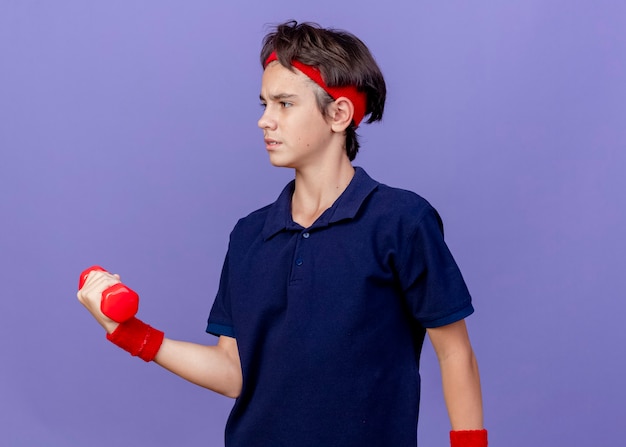 Stirnrunzelnder junger hübscher sportlicher Junge, der Stirnband und Armbänder mit Zahnspangen trägt, die Seite betrachten, die Hantel lokalisiert auf lila Wand hält