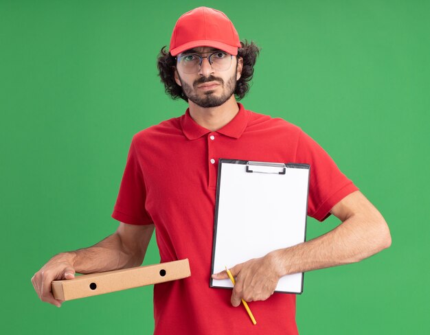 Kostenloses Foto stirnrunzelnder junger kaukasischer liefermann in roter uniform und mütze mit brille, der pizzapaket-bleistift hält und klemmbrett zur kamera zeigt, isoliert auf grüner wand