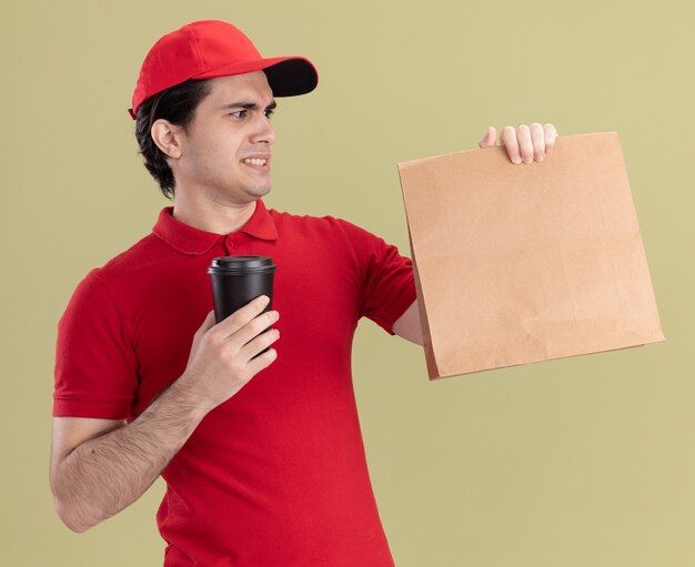 Stirnrunzelnder junger kaukasischer Liefermann in roter Uniform und Mütze mit Papierpaket und Plastikkaffeetasse mit Blick auf das Paket