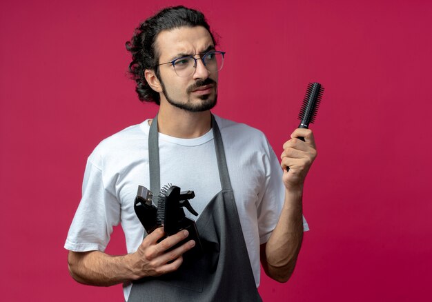 Stirnrunzelnder junger kaukasischer männlicher Friseur, der Brille und gewelltes Haarband in Uniform hält, die Haarschneidemaschine, Kamm, Sprühflasche hält Seite betrachtet auf purpurrotem Hintergrund mit Kopienraum