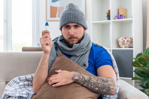 Stirnrunzelnder junger kranker Mann mit Schal und Wintermütze, der auf dem Sofa im Wohnzimmer sitzt und das Thermometer vertikal hält und das Kissen nach vorne schaut