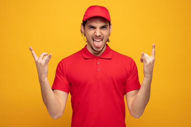 stirnrunzelnder junger lieferbote in uniform und kappe, der in die kamera blickt, die ein rockzeichen zeigt und die zunge isoliert auf gelbem hintergrund zeigt