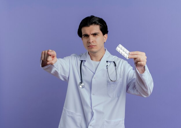 Stirnrunzelnder junger männlicher Arzt, der medizinische Robe und Stethoskop trägt, die Packung der Tabletten zeigt und lokalisiert auf lila Wand mit Kopienraum zeigt