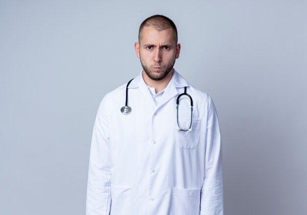 Stirnrunzelnder junger männlicher Arzt, der medizinische Robe und Stethoskop um seinen Hals trägt, der auf Weiß mit Kopienraum isoliert steht und schaut