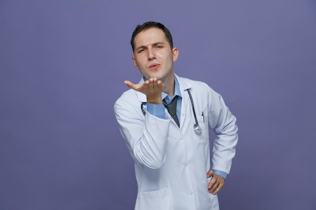 Stirnrunzelnder junger männlicher Arzt mit medizinischem Gewand und Stethoskop um den Hals, der die Hand auf der Taille hält und in die Kamera blickt, die einen Kuss isoliert auf violettem Hintergrund bläst