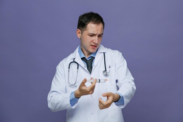 Stirnrunzelnder junger männlicher Arzt mit medizinischem Gewand und Stethoskop um den Hals, der die Hand in der Luft hält und eine Spritze mit Nadel hält, die isoliert auf violettem Hintergrund auf seinen Arm blickt