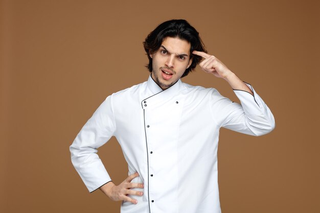 Stirnrunzelnder junger männlicher Koch in Uniform, der die Hand auf der Taille hält und in die Kamera blickt und eine Denkgeste zeigt, die auf braunem Hintergrund isoliert ist