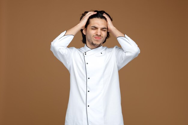 Stirnrunzelnder junger männlicher Koch in Uniform, der mit geschlossenen Augen die Hände auf dem Kopf hält und Kopfschmerzen isoliert auf braunem Hintergrund hat