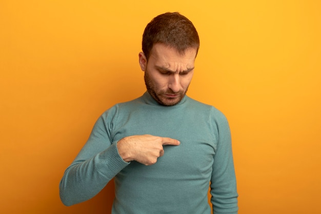 Stirnrunzelnder junger Mann, der Finger auf Brust zeigt, betrachtet Brust lokalisiert auf orange Wand
