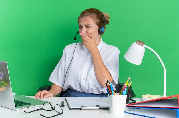 Stirnrunzelndes junges blondes Call-Center-Mädchen mit Headset am Schreibtisch sitzend mit Arbeitswerkzeugen, die die Hand auf dem Mund halten und auf den Laptop schauen, der auf grüner Wand isoliert ist