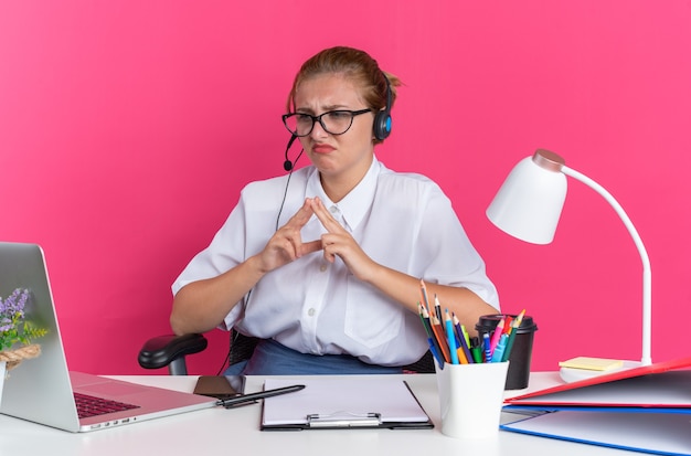 Stirnrunzelndes junges blondes Callcenter-Mädchen mit Headset und Brille, das am Schreibtisch mit Arbeitswerkzeugen sitzt und die Hände zusammenhält und den Laptop isoliert auf rosa Wand betrachtet