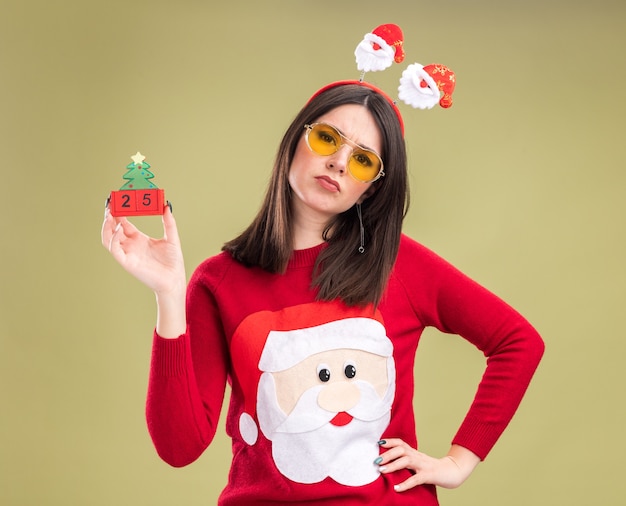 Stirnrunzelndes junges hübsches kaukasisches Mädchen, das Weihnachtsmann-Pullover und Stirnband mit Brille trägt, die Weihnachtsbaumspielzeug mit datumshaltender Hand halten