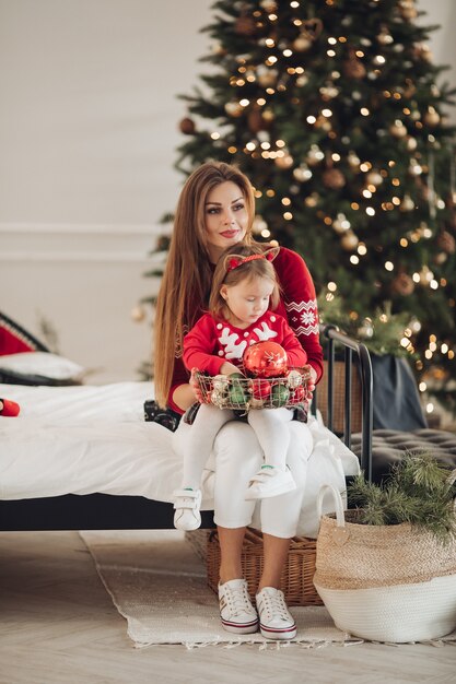 Stock Foto der liebenden Mutter im grünen Kleid, das ihrer kleinen Tochter im Pyjamakleid ein Weihnachtsgeschenk gibt. Sie stehen neben einem wunderschön geschmückten Weihnachtsbaum unter Schneefall.