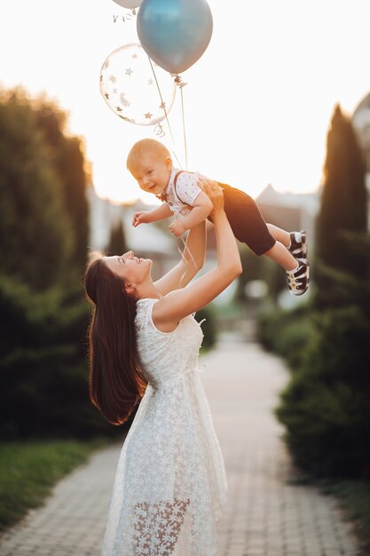 Stock Foto einer liebevollen Mutter im schönen weißen Sommerkleid, die ihren Sohn mit aufblasbaren Luftballons in der Luft im schönen Garten in unscharfem Hintergrund aufzieht. Geburtstag des Sohnes im Freien feiern.