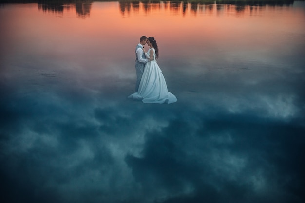 Stock Foto einer romantischen Braut im Hochzeitskleid und Bräutigam im Anzug umarmt von Angesicht zu Angesicht stehen auf nassem Sand mit Himmelsreflexion darauf. Wolken, die auf dem Boden reflektieren, machen eine fantastische Aussicht.