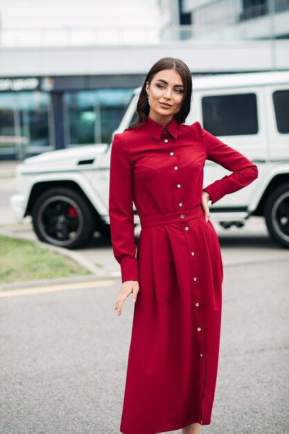 Stock Foto einer schönen jungen Dame in leuchtend rotem Kleid mit braunen Haaren und Make-up lächelnd, während sie mit der Hand auf der Taille auf der Straße posiert.