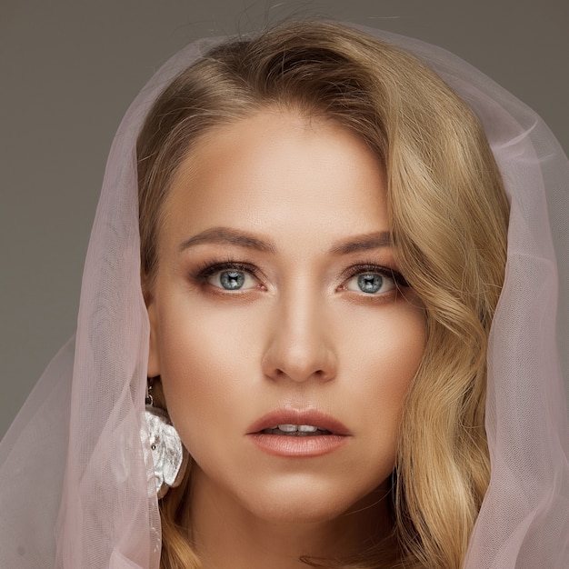 Stock Foto einer wunderschönen blonden Frau mit blauen Augen und natürlichem Make-up, die einen rosa Schleier trägt und eine zerbrechliche rosa Rose hält. Braut-Konzept.