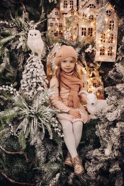 Stock Foto von entzückenden brünetten Mädchen mit Pfirsich Strickmütze und Schal sitzen in Weihnachtsschmuck mit weißen Spielzeugkaninchen und Eule. Beleuchtete Häuser im Hintergrund.