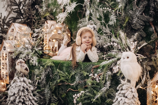Stock Foto von entzückenden kleinen kaukasischen Kind in beige Ohrenschützer Verlegung mit Kinn auf Händen, umgeben von Weihnachtsschmuck und lächelnd in die Kamera. Winterwunderland-Konzept.