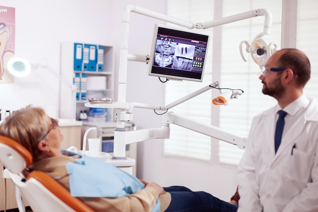 Stomatolog spricht über zahnbehandlung mit einer älteren frau in der zahnarztpraxis. medizinischer zahnpfleger, der auf die röntgenaufnahme des patienten auf dem bildschirm zeigt, der auf einem stuhl sitzt.