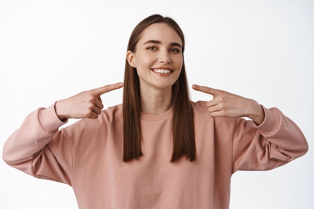 Stomatologie und Zahnklinikkonzept. Lächelnde Frau, die auf ihr perfektes weißes Lächeln mit zufriedenem Gesicht zeigt und Zahnarzt empfiehlt, weißer Hintergrund