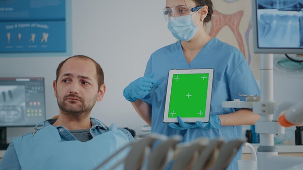 Stomatologiekrankenschwester, die Tablette mit grünem Bildschirm vertikal hält, den isolierten Kopienraum mit Chroma-Key-Hintergrund erklärt und dem Patienten bei der Mundpflegeuntersuchung eine Vorlage nachahmt.