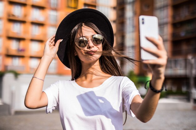 Straße im Freien der jungen trendigen Frau, die Selfie auf der Straße macht und stilvollen Hipsterhut trägt.