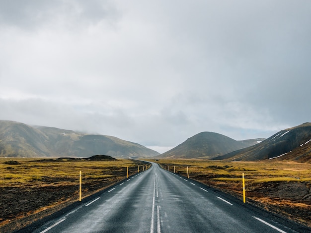 Straße umgeben von Hügeln, die mit Grün und Schnee unter einem bewölkten Himmel in Island bedeckt sind