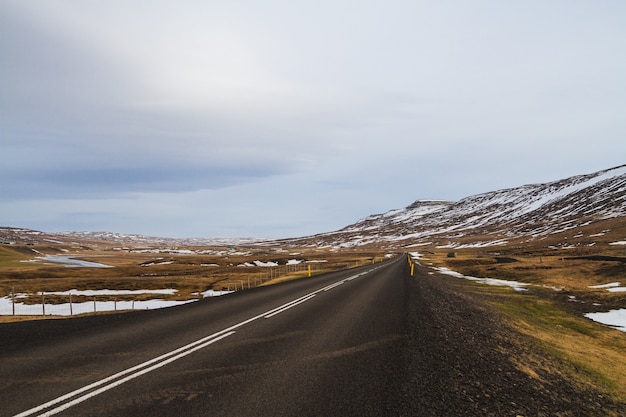 Straße umgeben von Hügeln, die mit Schnee und Grün unter einem bewölkten Himmel in Island bedeckt sind