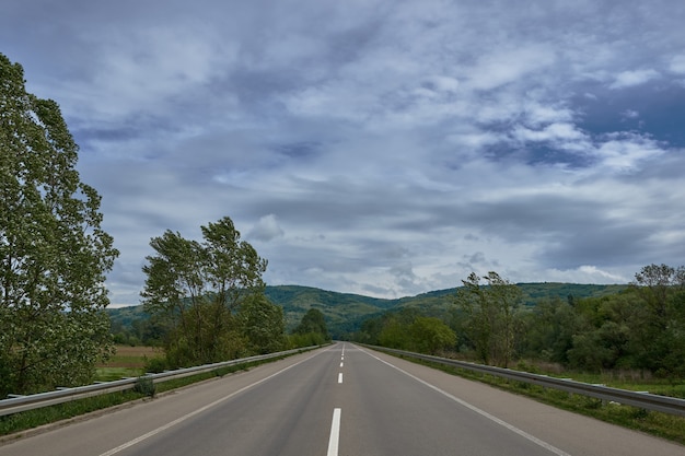 Straße umgeben von Hügeln, die tagsüber unter dem bewölkten Himmel mit Wäldern bedeckt sind