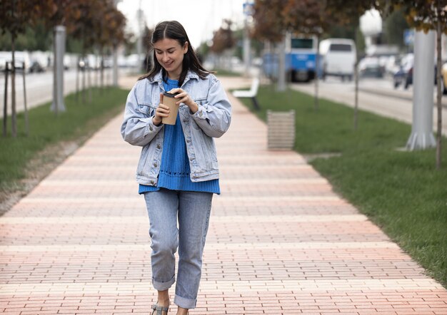 Straßenfoto einer attraktiven jungen Frau, die mit einem Kaffee in der Hand eine Stadtstraße entlang geht