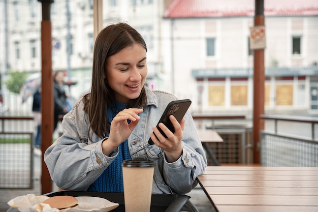 Straßenporträt einer fröhlichen jungen Frau auf einer Café-Terrasse, die ein Telefon hält