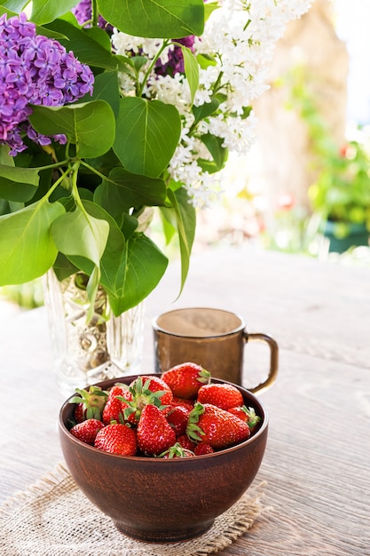 Strauß lila Zweige in Kristallvase, Tonschale mit roter Erdbeere und dunkler Glasschale auf Holztisch.