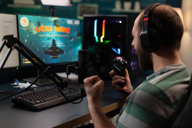 Streamer gewinnende videospiele mit joystick auf dem computer. man live-streaming-gameplay und feiert den sieg, indem er mikrofon und kopfhörer verwendet, während er mit dem online-chat auf dem monitor spricht. spieler im stream