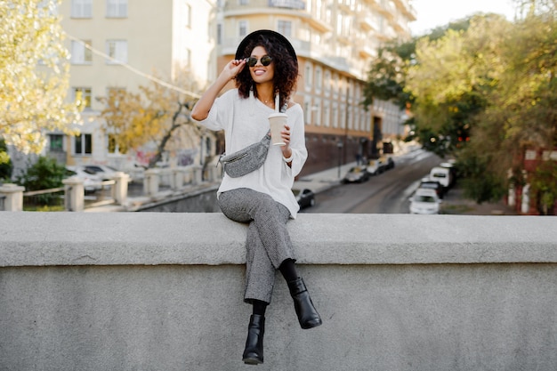 Street Fashion Look. Stilvolles schwarzes Mädchen, das auf der Brücke sitzt und während ihrer Freizeit Tasse Kaffee oder Tee hält. Freiberufliche Frau. Tragen eines schwarzen Hutes und einer Sonnenbrille.