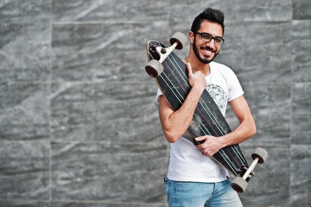 Streetstyle-arabischer Mann mit Brille und Longboard posierte vor grauer Wand