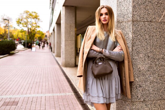 Streetstyle-Modeporträt der eleganten glamourösen blonden Frau, die auf der europäischen Straße nahe Einkaufszentrum, elegantem Kleidpullover und Kaschmirmantel, getönten Farben, Frühlingszeit aufwirft.