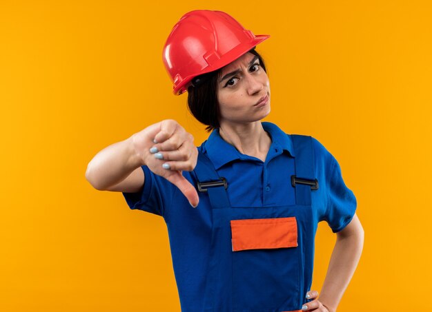 Strenge junge Baumeisterin in Uniform, die den Daumen nach unten zeigt und die Hand auf die Hüfte legt, isoliert auf gelber Wand